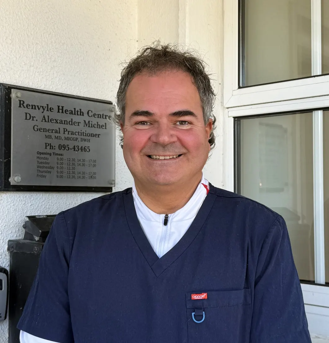 Dr Alexander Michel, smiling, at the entrance of Renvyle Health Centre beside the practice nameplate.