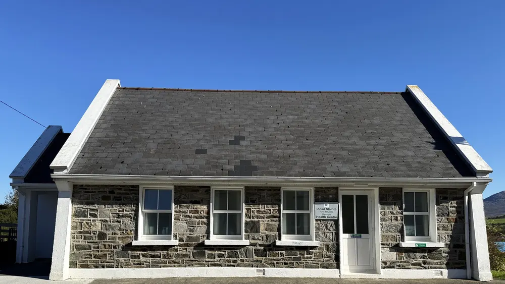 Renvyle Health Centre in Tully: the stone-fronted building with its slate roof, white gables, white door and sash windows, under a clear blue sky.